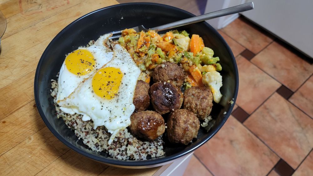 Black dish with quinoa, mixed (formerly frozen) veggies, meatballs (also formerly frozen), and two sunny side up eggs. Less visible is the barbecue sauce drizzled over the meatballs and veg 