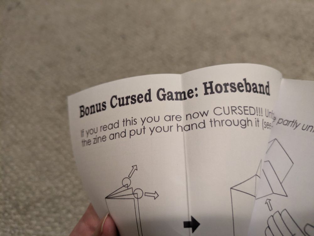 A hand holding a piece of paper that says "Bonus Cursed Game: Horse and. If you read this you are now CURSED!!!" Some text is cut off and the next line says "the zine and put your hand through it". Below the text on the paper are illustrations on how to handle the zine, mostly cut off in the picture