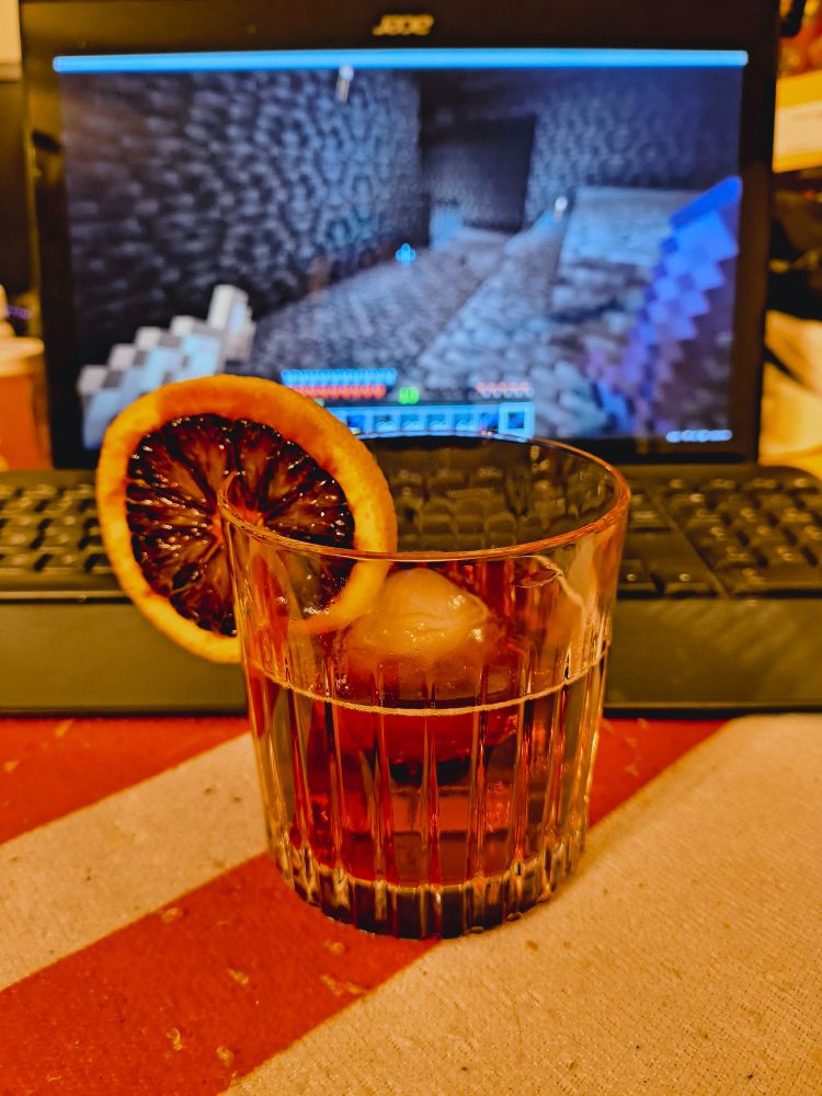 An Old Pal cocktail in a rocks glass with a single rough spheroid ice cube and a blood orange wheel garnish in front of a keyboard and monitor. Minecraft is shown on the monitor.