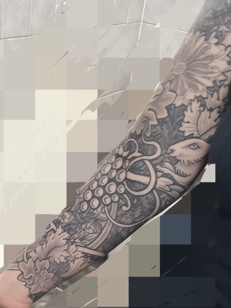 A white persons arm with William Morris black and gray design tattoo covering it. It has a rabbit, some grapes, a vine and a dark background print of oak and thistle leaves. 