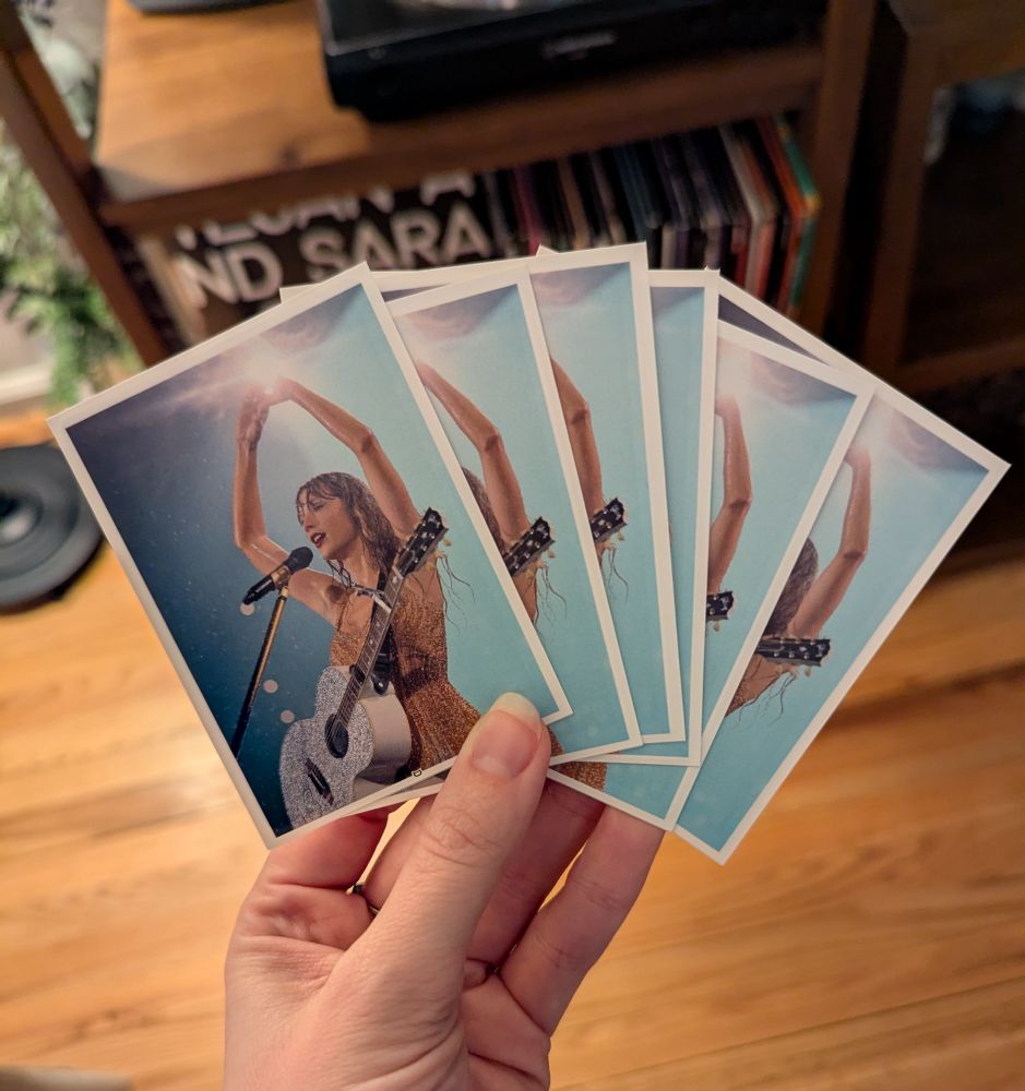 My hand holding six cards that are sleeved, and the sleeve backs are the quoted image of a rain soaked Taylor Swift during the Fearless set of the Eras Tour, heart hands in the air