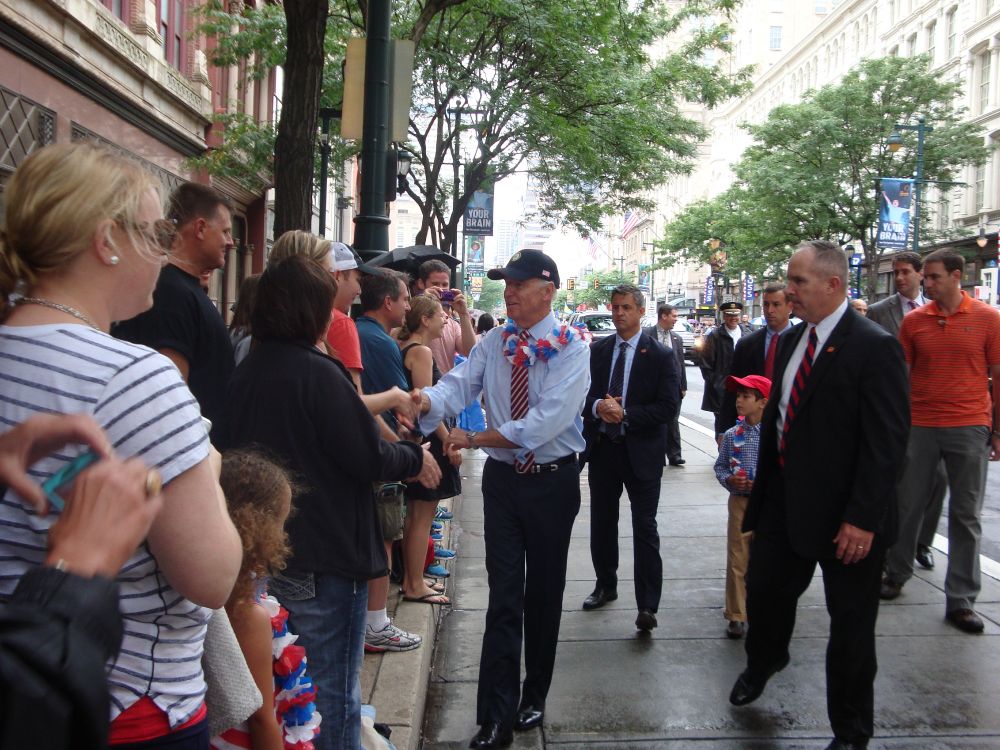 Joe Biden walking along a sidewalk, shaking the hands of spectators in the 2014 Fourth of July parade in downtown Philadelphia