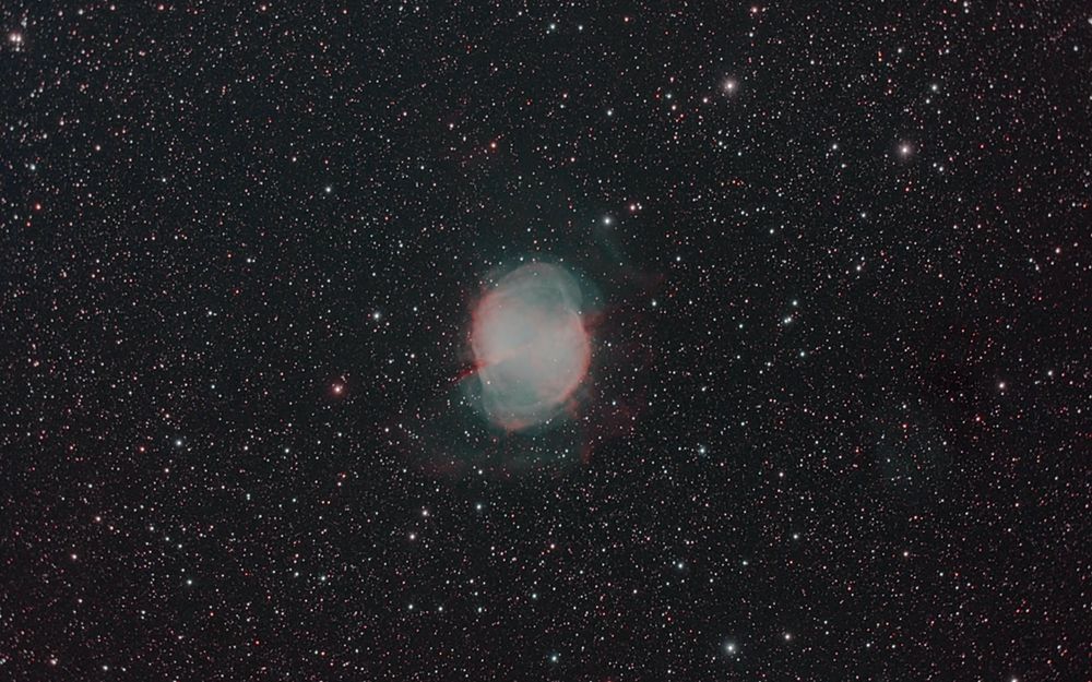 A planentary nebula, Messier 27 fills the center tenth to an eighth of the field of view, surrounded by a dense starfield against a black background. The nebula is mostly blue-green (from the O-3 emission) but there is some pink from hydrogen emission. The famous dumbbell shape is formed left-right in the image, with the outer edges of the blue-green dumbbell (which is the brightest part of the nebula) tinged pink. An X of pink nodules crosses the bright dumbbell part. Above and below the dumbbell, filling in the missing space to make the nebula more circular, is lacy blue-greenish nebula. The bottom edge is, again, tinged pink. A very faint green and pink arc surrounds the bright nebula at center, the result of an earlier outgassing from the central star. 

The image was made with a Celestron 8 Schmidt Cassegrain working at f/7, a ZWO ASI2600MC camera, and an Optolong L-Quad light pollution filter from within the city limits of Winston-Salem, NC. The image is the result of a stack of 90 5 minute exposures processed in Astro Pixel Processor and Photoshop. No AI was involved, it's all real data. 