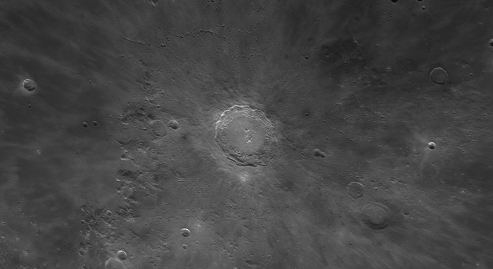 Crater Copernicus near local noon so that there are few shadows and little relief. Hundreds of tiny craterlets ring the land surrounding the crater and the ray system from the ancient impact is clearly seen as ghostly lighter material splashed across the darker maria. Only a few moderately sized craters share the frame with the large, bright Copernicus at center. To the upper left, near the edge, is Pytheas, about a quarter the size of Copernicus. To the right of center is the double crater Fauth arcing over to Rheinhold at lower right. Copernicus itself is very bright under the high Sun, with it’s myriad central peaks bright specks against the rough crater floor. Only by looking at the terraced walls can we see that the Sun is a little past noon – the upper rim is extremely bright, while there is just a hint of a shadow on the first terrace on the bottom. 