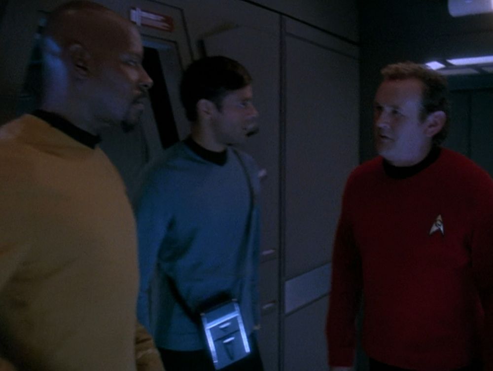 Three men stand in a dimly lit narrow corridor of the starship Defiant talking. They are wearing gold, blue, and red tunics.