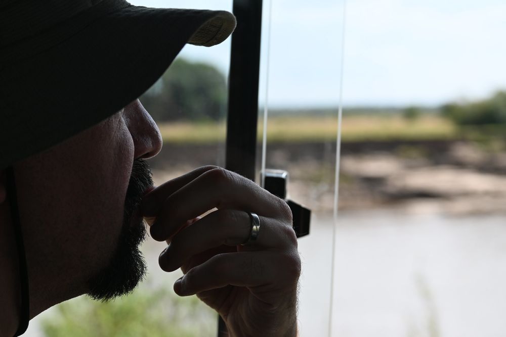 a bearded white man wearing a wide-brimmed hat and a ring, looking out a window to an out-of-focus scene of a riverbank