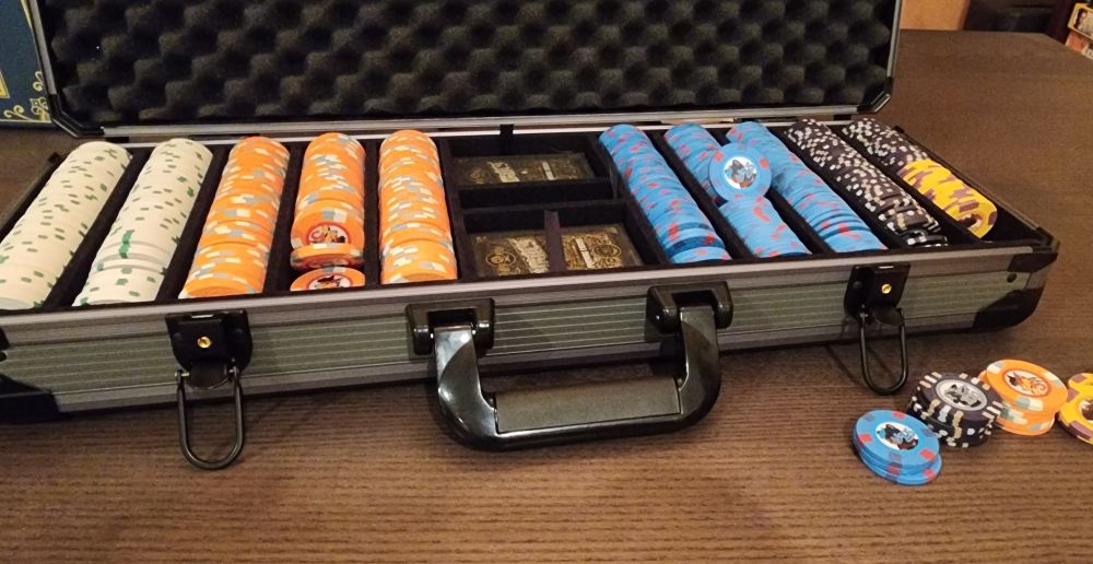 A photograph of the poker chip set in its padded latching carrying case on a wooden table. There are ten rows of chips in five different denominations. There are two decks of cards in the case.