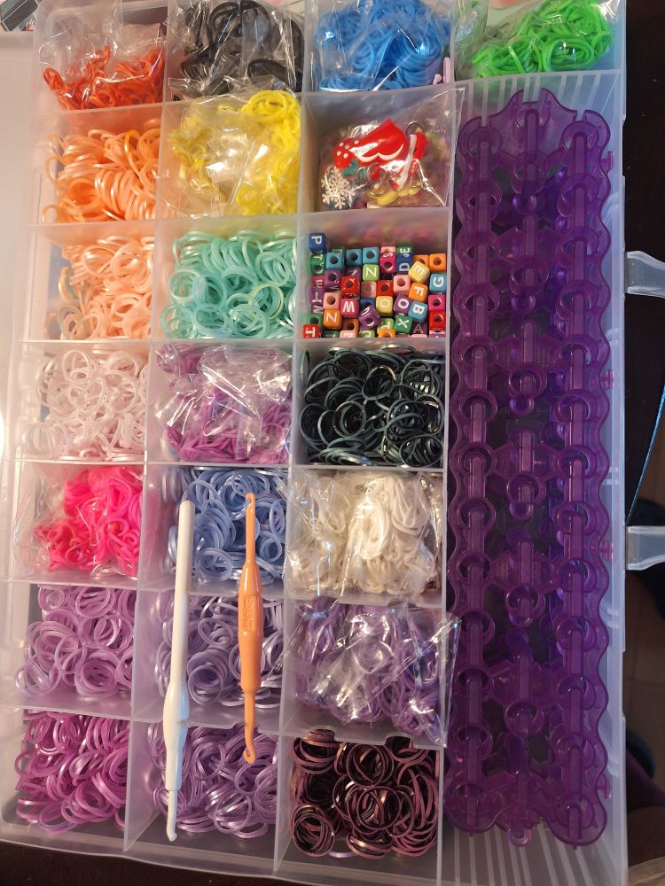 A tray with dividers filled with rubber bands of various colors and some assorted supplies. So I used to do a lot of rainbow loom. I made pencil huggers, bracelets, keychains, etc. This is what I used to do that. Rubber bands, a loom, hooks, beads, and some charms