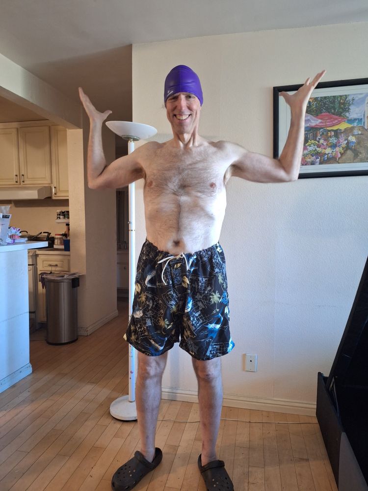 A skinny dude in swim trunks and Crocs make a sort of shrug pose. He looks ridiculous in a purple swim cap.