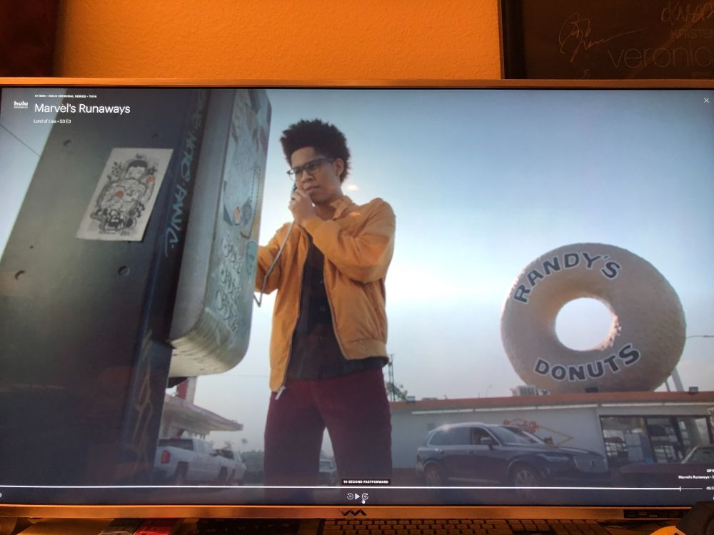 Screenshot from Marvel's Runway, with a character in the foreground and Randy's Donuts in the background.