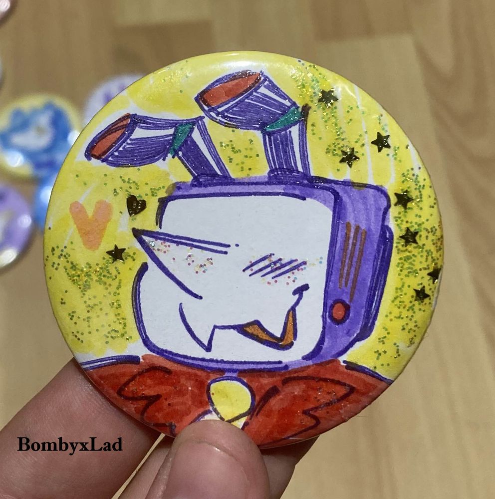 [ID: A pin button drawn in markers. The design is a smiling TV head person with chunky antennae, surrounded by starry glitter. end ID]

