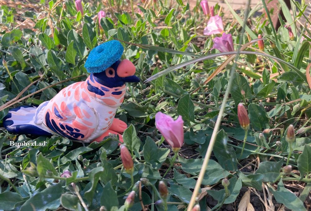 [ID: this pigeon sculpture has a tiny beret. it sits in green leaves and pink flowers. end ID]
