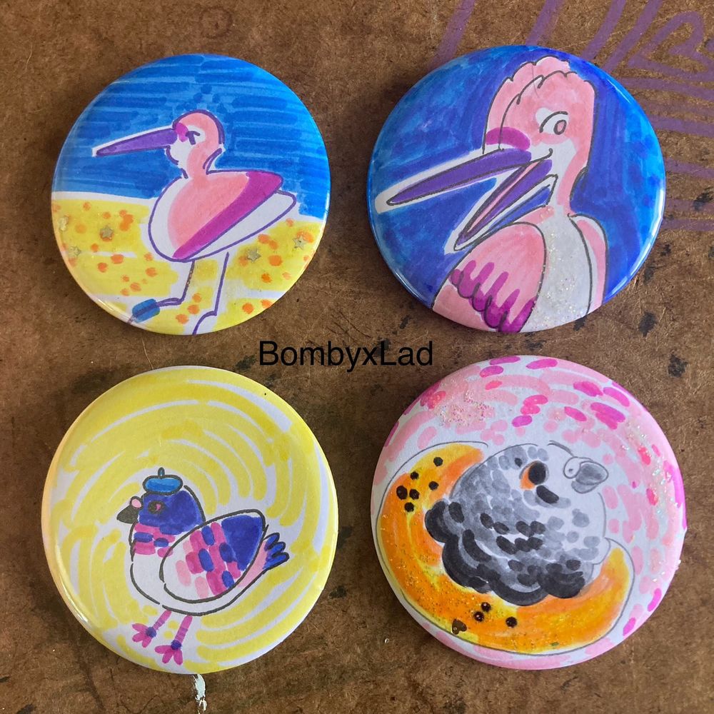 [Four birds on pin buttons, drawn in marker. Two are of a pink shorebird, one is a mottled pink pigeon in a beret, and one is a mottled grey pigeon wearing a bagel around its neck. end ID] 