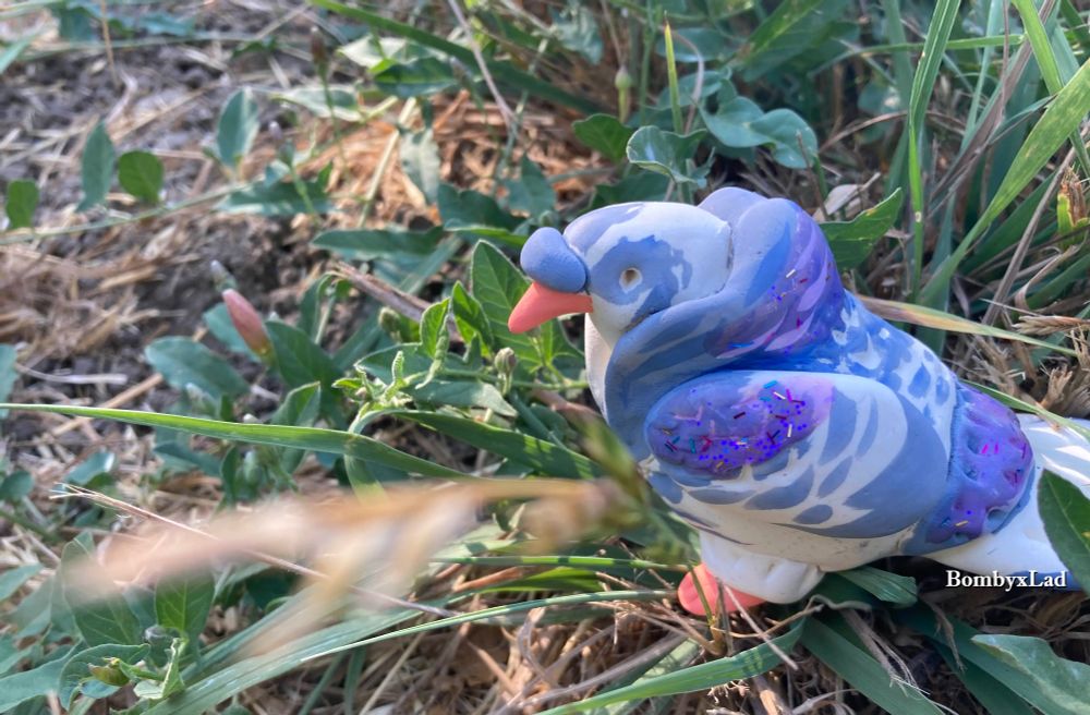[ID: sculpture of a pigeon sitting amongst real leaves and grass. It has motled patterns in grey and white. end ID]
