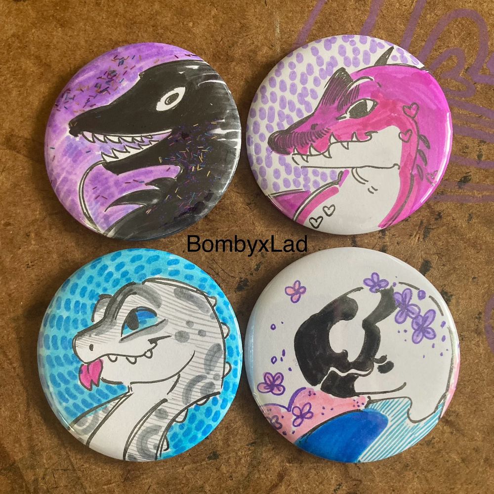 [ID: Four reptalian creatures on pin buttons, drawn in marker. A fluffy black worm, pink spinosaurus, grey leopard gecko, and mottled monster with flower crown. end ID]
