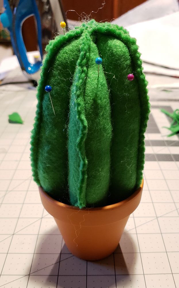 A pincushion shaped like a cactus. It sits in a tiny terra cotta pot.