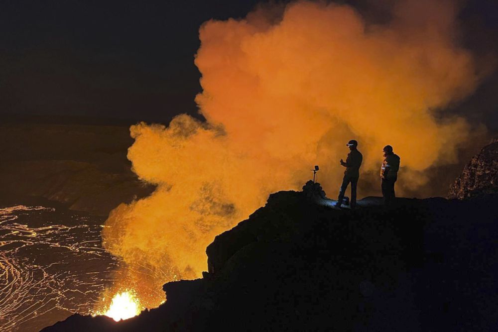 Lava shoots high into the sky. Molten rock erupts from two vents simultaneously. The nighttime sky glows red and orange, reflecting the lava oozing across a summit crater.