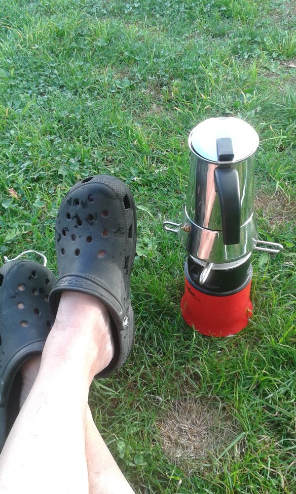 Roter Campinggasbrenner mit Bialetti-Espressokocher auf grüner Wiese. Davor wartend, weisse Beine in Crocs.