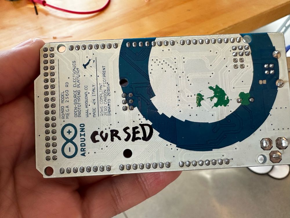 Photo: the back side of an Arduino Mega board. Written in trembling Sharpie at the bottom left: “Cursed” in all caps.