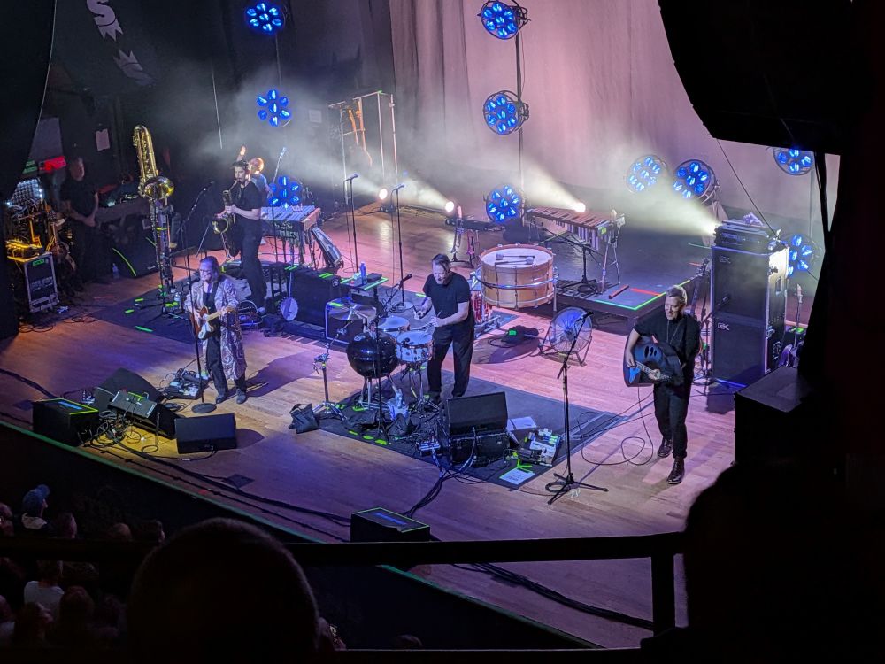Violent Femmes play on stage. They have a number of strange instruments including a comically shaped bass, a barbeque grill in the drum set, and a massive bass drum.