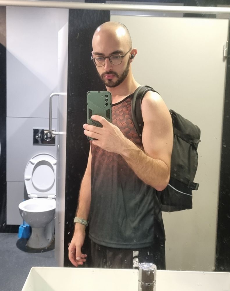 Foto do espelho da academia, na frente de uma pia e cabines de vaso sanitário. Eu sou careca e branco, estou de óculos, camiseta e short de malhar, e uma mochila nas costas. 