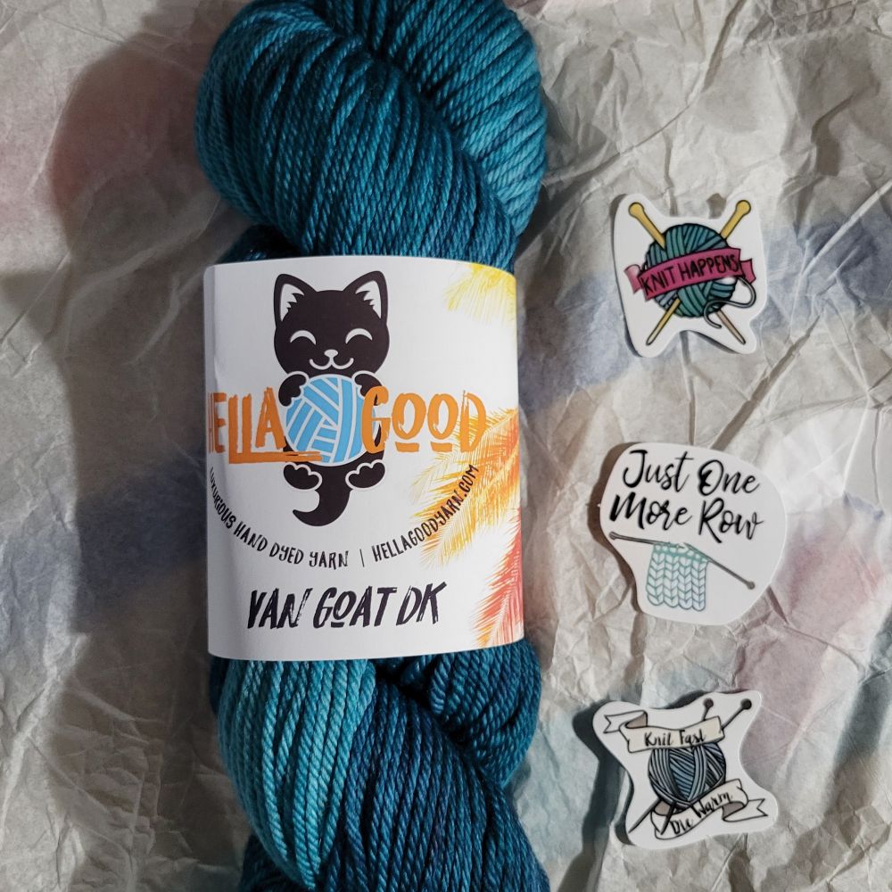 Blue variegated yarn wrapped in a label with a cartoon black cat printed on it. The yarn is laying on white tissue paper and there are three knitting themed stickers next to it.