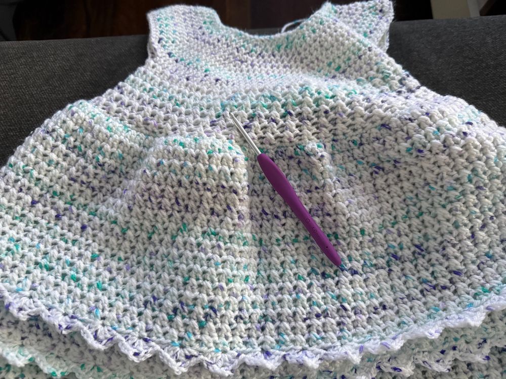 a crocheted baby dress that is white with green and purple speckles plus a purple crochet hook
