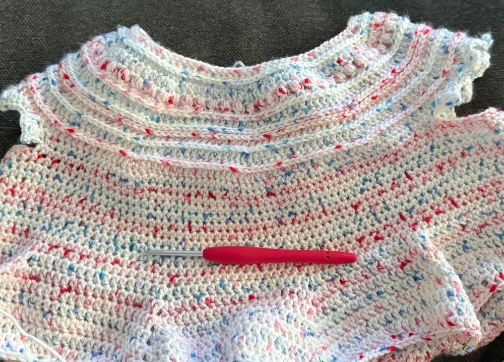 a crocheted item that is white with red and blue speckles plus a red crochet hook