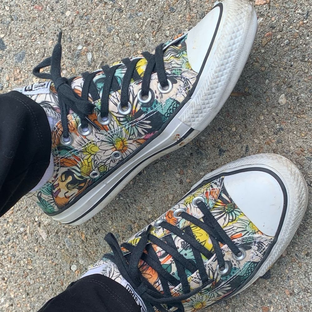 My feet on concrete pavement in low top chuck Taylor sneakers. There is a floral pattern in black outline with splotches of teal, yellow, and orange, plus black laces. 