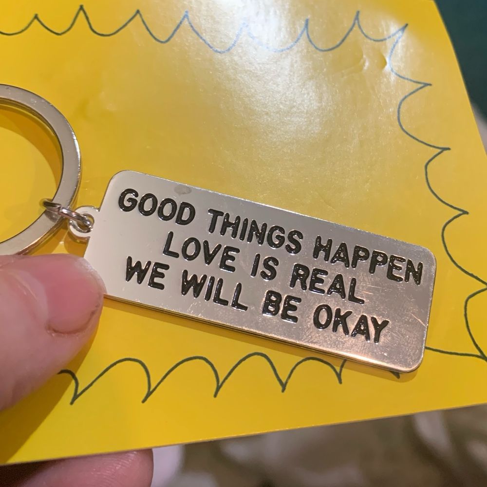 Photo of a metal keychain still attached to its packaging which reads "good things happen love is real we will be OK" 