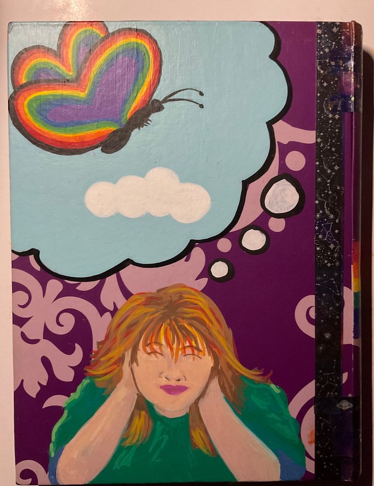 A hand-painted book cover against a purple patterned background. At the bottom, a portrait depicts a person with reddish-blonde hair covering their ears with their hands. A large, light-blue thought bubble emerges from their head, containing a butterfly with heart-shaped rainbow wings. The book's spine on the right is decorated with black constellation-themed tape.

