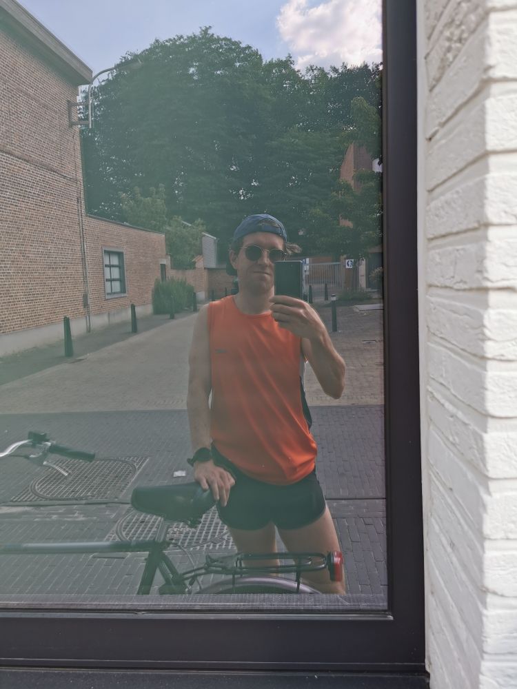 Selfie van Pieter Van der Schoot in hardloopoutfit in spiegelbeeld in een raam. Hij draagt een blauwe pet achterstevoren, een zonnebril van Ray Ban, een oranje mouwloos shirt en een zwarte loopshirt. 