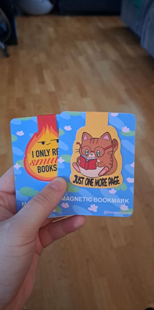 Two bookmarks attached to a blue, river like backingboard with waterlillies. One bookmark is an orange cat reading a book with the text "just one more page" with a yellow background. The other is a sassy flame that says "i only read smutty books". 