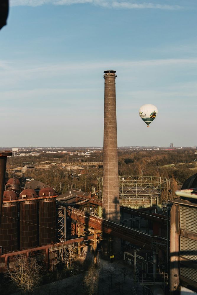 Ein Blick über das Ruhrgebiet. Im Vordergrund der Schornstein eines Hochofens. Am blaugrauen Himmel ein Heißluftballon.