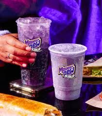 A promotional photo for Mountain Dew Baja Midnight. It's a purple drink in soda and freeze form. Tacos and burritos and a disembodied hand herald its arrival. 