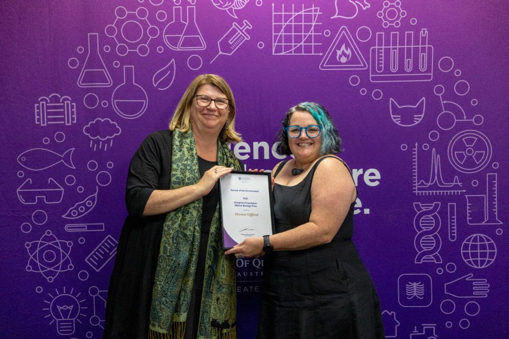 Two women in black standing in front of a purple background holding a framed award.