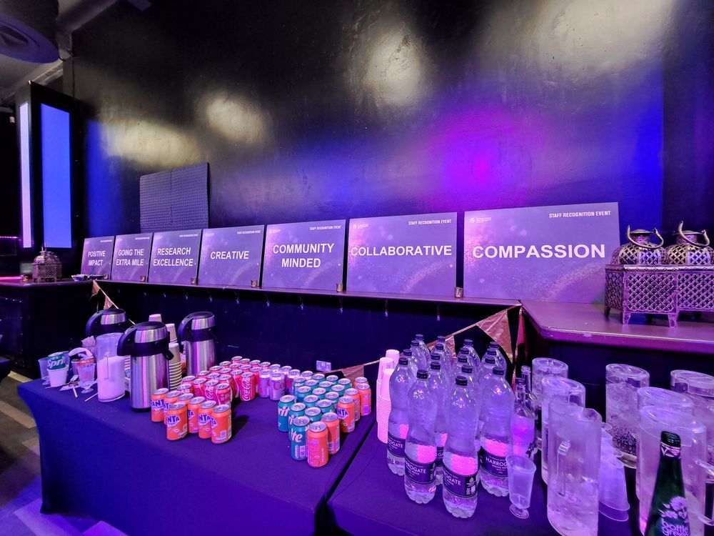 A long table laden with drinks and refreshments at a "Staff Recognition Event." On the table are various bottled waters, cans of soda, and insulated dispensers. In the background, several illuminated signs are visible, displaying words like "POSITIVE IMPACT," "DOING THE EXTRA MILE," "RESEARCH EXCELLENCE," "CREATIVE," "COMMUNITY MINDED," "COLLABORATIVE," and "COMPASSION."