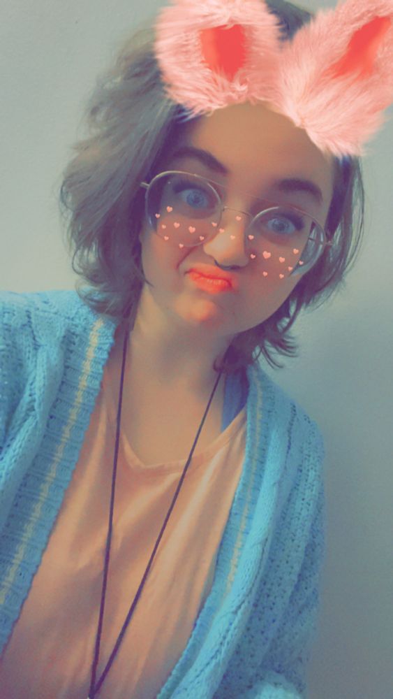 a girl with a bunny filter wears a necklace and makes a funny face at the camera. she wears a light blue cardigan and pink shirt 