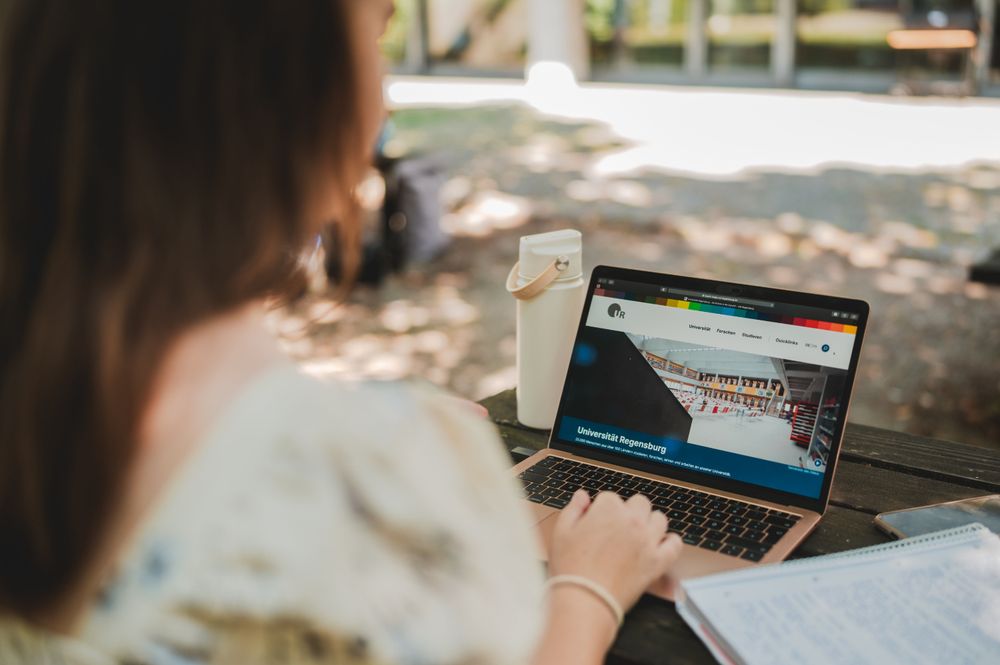 Auf der linken Seite sitzt eine junge Frau im Freien mit dem Rücken zur Kamera. Im Zentrum des Bildes steht ein Laptop, auf dem Bildschirm ist die neue Seite der Universität Regensburg geöffnet. Daneben steht eine Trinkflasche.