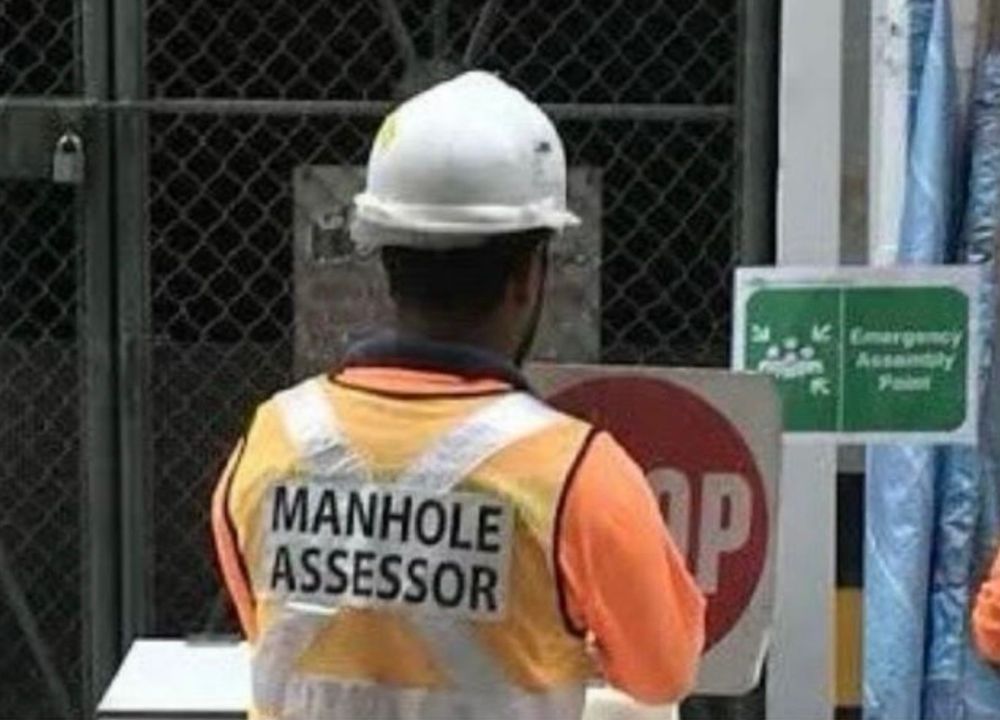 View from behind of a guy in a hard hat and hi-viz vest that has "Manhole Assessor" written on the back