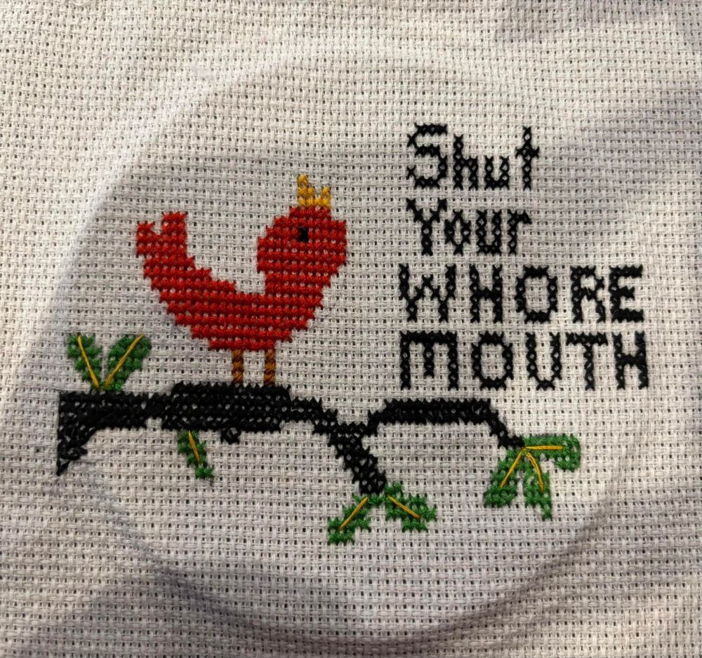A cross-stitch pattern of a badly drawn red bird wearing a crown standing on a branch.
Text is:
"Shut your
whore mouth"

(I don't possess this item.)