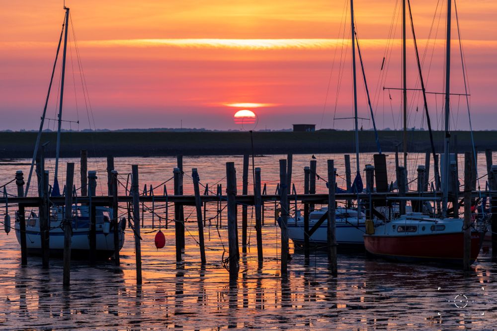 Segelboote im Hafen bei Ebbe, davor ein Holzsteg. Über dem Horizont geht die Sonne auf und taucht den Himmel und das Wasser in orange-rote Farben.