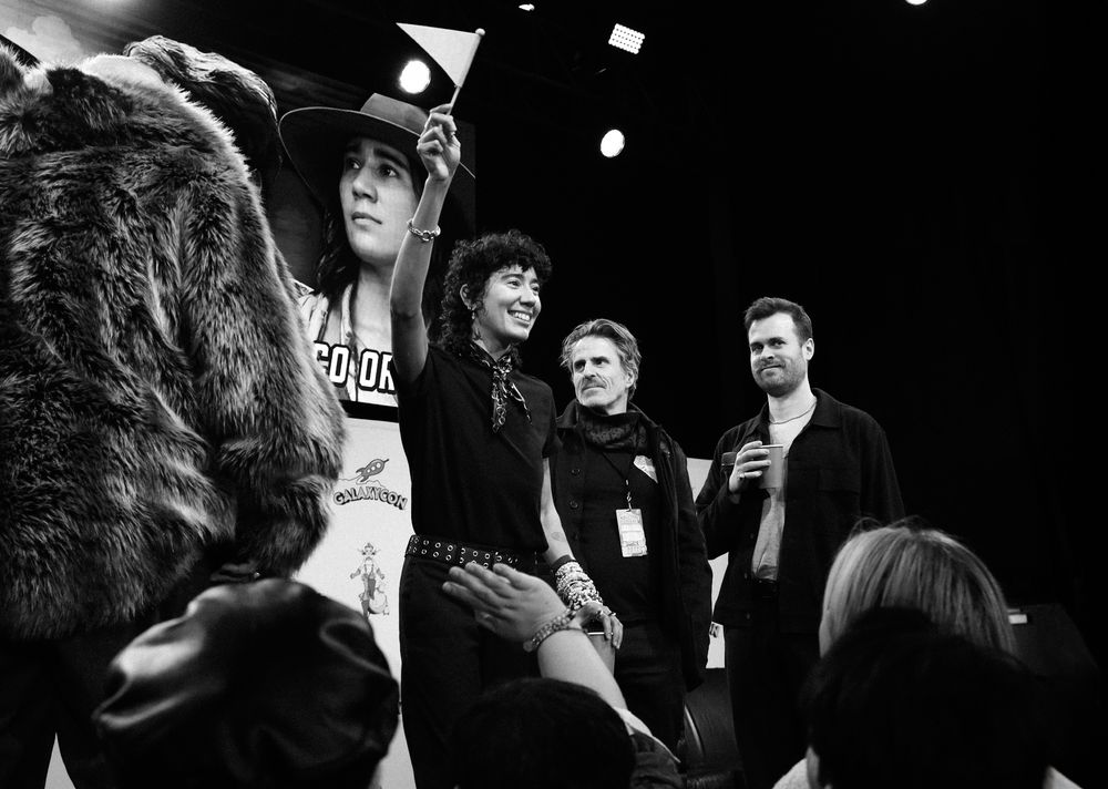 Black and white photograph of Vico Ortiz, Con O’Neill, and Nathan Foad onstage at the Our Flag Means Death panel at GalaxyCon Columbus. Kristian Nairn is in the corner of the photograph with his back to the crowd, and Vico Ortiz is waving a flag that says “SLUTS” on it. 