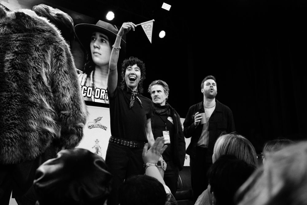 Black and white photograph of Vico Ortiz, Con O’Neill, and Nathan Foad onstage at the Our Flag Means Death panel at GalaxyCon Columbus. Kristian Nairn is in the corner of the photograph with his back to the crowd, and Vico Ortiz is waving a flag that says “SLUTS” on it. Con O’Neill is looking off to the side handsomely while Nathan Foad is making a silly face at the crowd. 