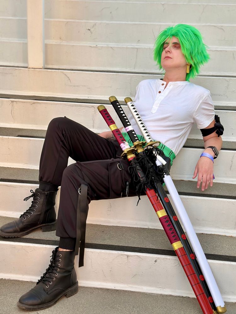 Person dressed as Zoro from one piece - they have green hair and are wearing a white t shirt, black pants, and black boots. The person’s eyes are closed as they sit leaning back on a set of stairs