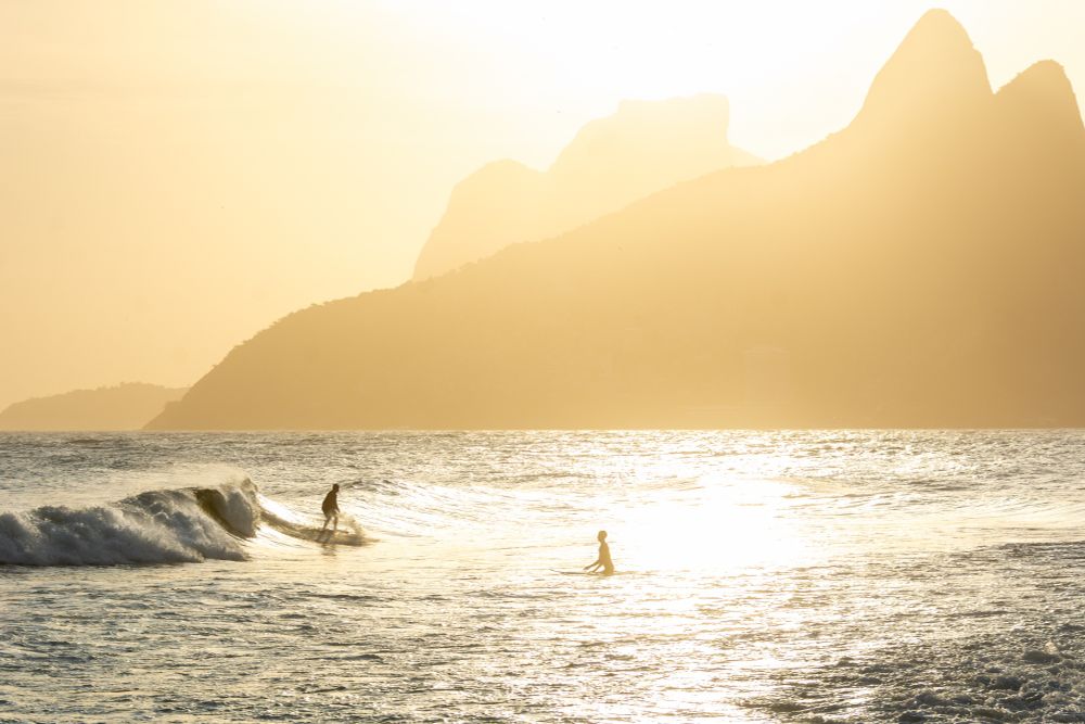 A golden sunset over the iconic hills near Ipanema Beach in Rio de Janeiro with two surfers in the water 