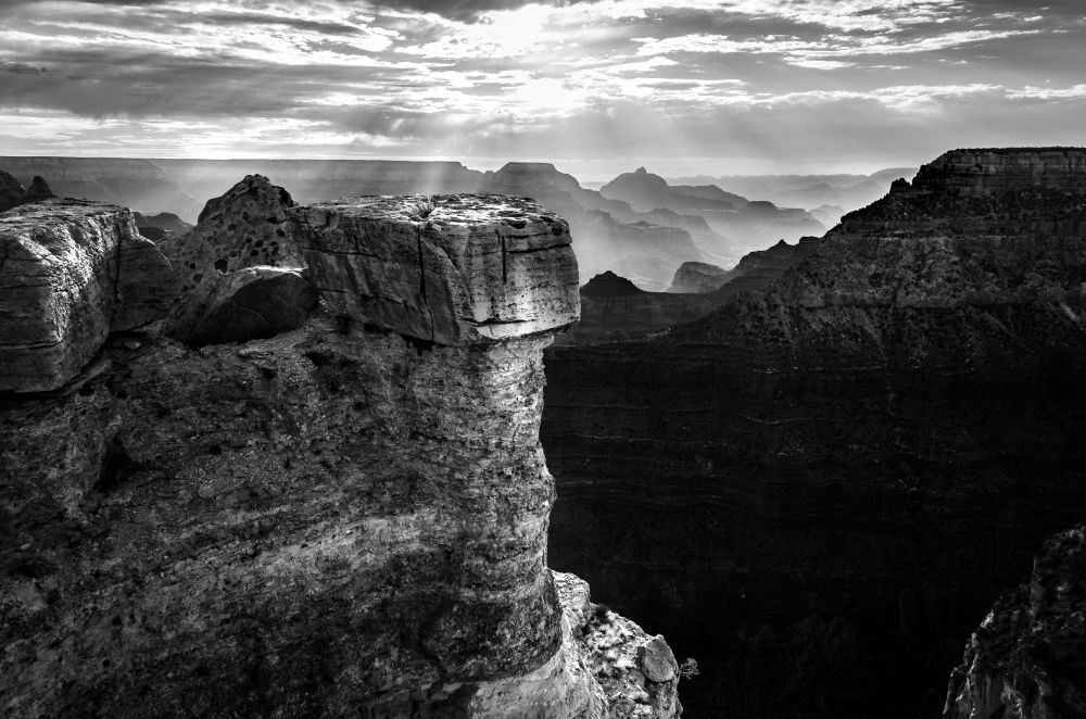 Black and white shot of the sunrise over the Grand Canyon with a scenic cliff ledge in the foreground