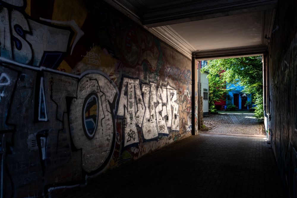 Foto eines Tunnels mit Graffitis an den Wänden, der zu einem Hinterhof mit Bäumen und bunt gestrichenen Hausfassaden führt 