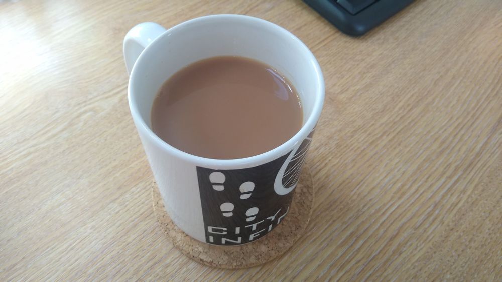 A City Infinity branded mug sitting on a cork coaster on a light wooden desk with a keyboard just out of shot.