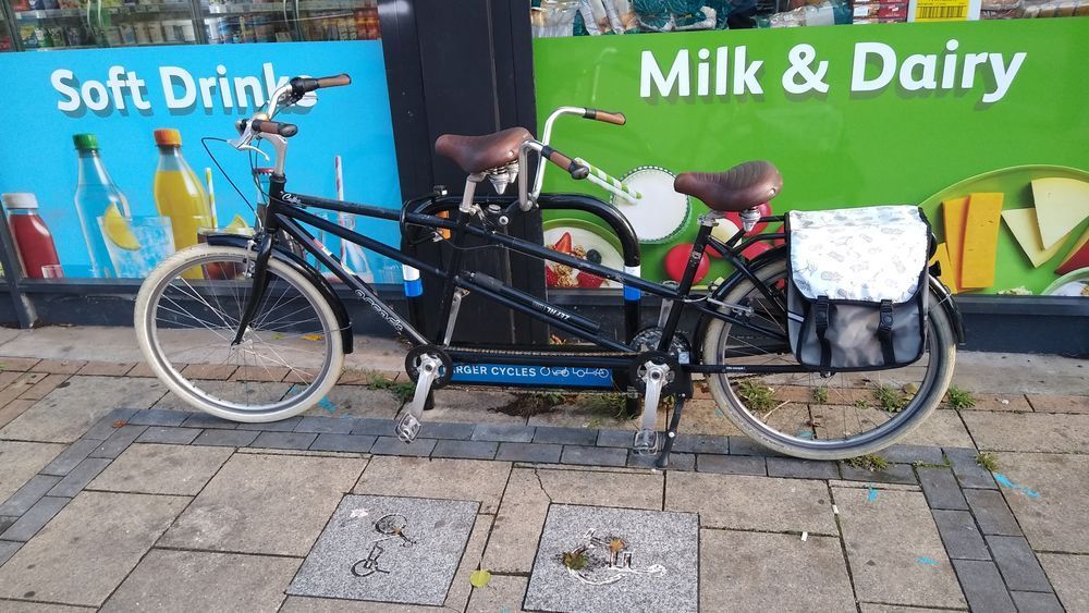 A black tandem bicycle with chrome handlebars and brown seats locked to a cycle parking hoop reserved for non-standard cycles. The paving is light slabs with a dark surround to mark the area and there are two other slabs, one showing a cargocycle and one showing the Disabled person logo.